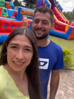 Selfie de um casal sorrindo em frente aos brinquedos infláveis da Miranda Park montados em um gramado